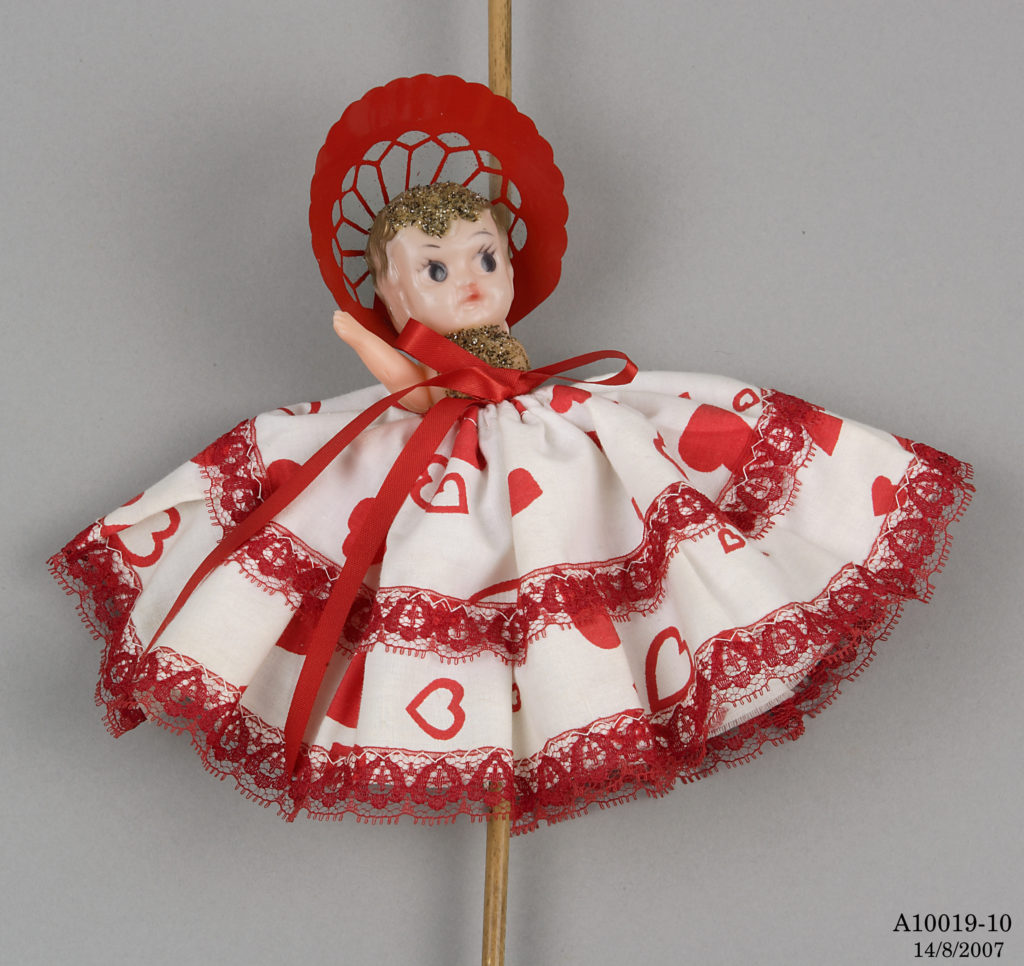 Small plastic doll with gold painted hair wearing a red bonnet and a sequin top. She is dressed in a full skirt made of white fabric decorated with red hearts and red lace and ribbons. The doll is attached to a cane walking stick. 